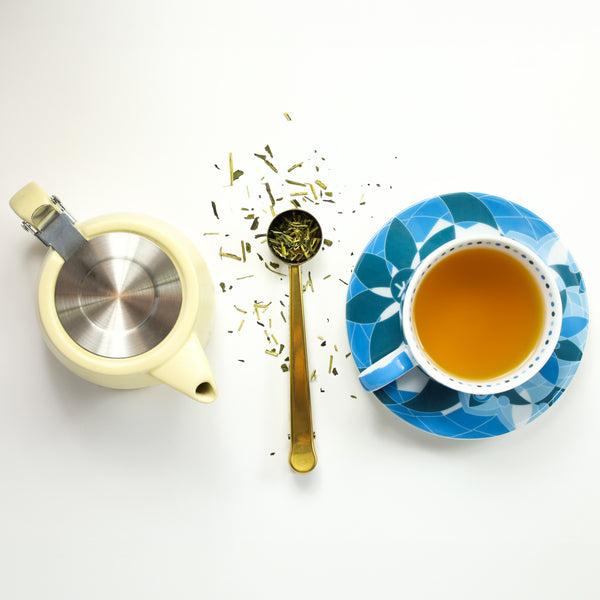 Cup & Saucer filled with brewed Kukicha tea next to Gold Tea Scoop and Nordic Tea Pot.