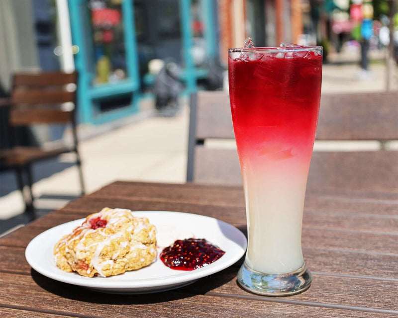 Outdoors a layered Raspberry Lavender lemonade in a tall glass next to a strawberry scone