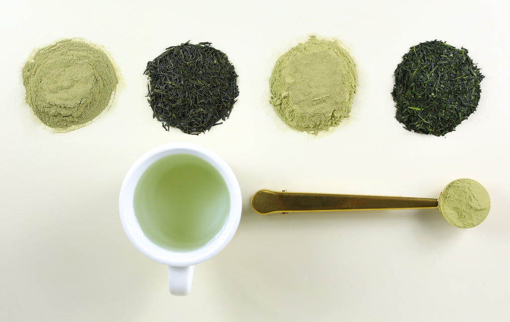 Piles of matcha powder and loose leaf green tea next to tea cup with green tea and gold tea scoop with matcha powder