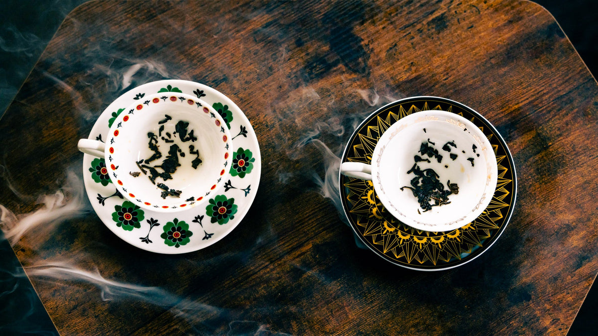 Overhead view of two patterned teacups with mysterious tea leaf formations, as ghostly wisps of smoke swirl across a dark wooden table.
