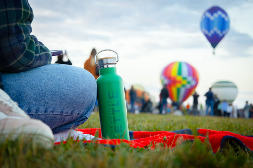 A green Whistling Kettle insulated tea flask on a picnic blanket during a hot air balloon festival