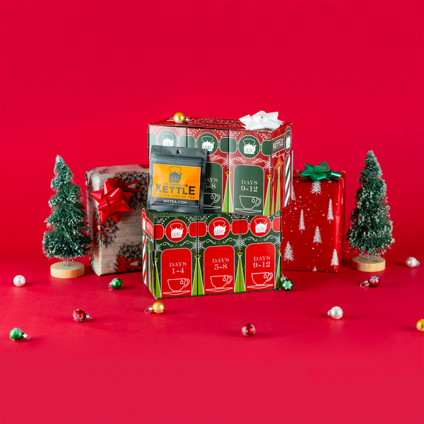 Two 12 Days of Tea advent calendar boxes are stacked among wrapped gifts, miniature Christmas trees, bows, and ornaments on a festive red background