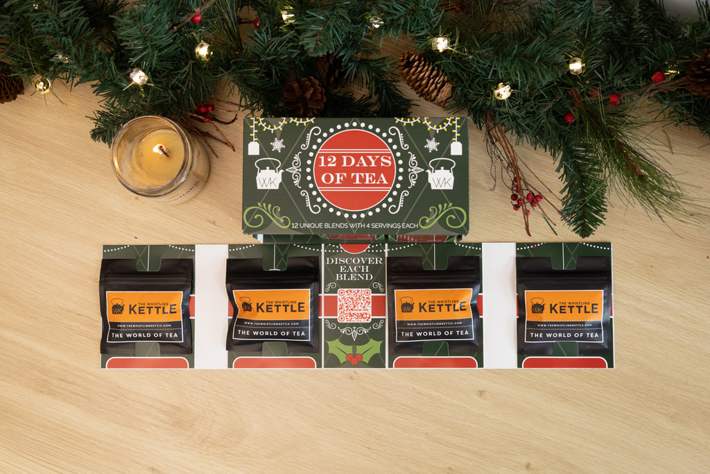 Overhead view of an open 12 Days of Tea advent calendar displaying individual tea packets beneath Christmas greenery, pinecones, lights, berries, and a glowing candle