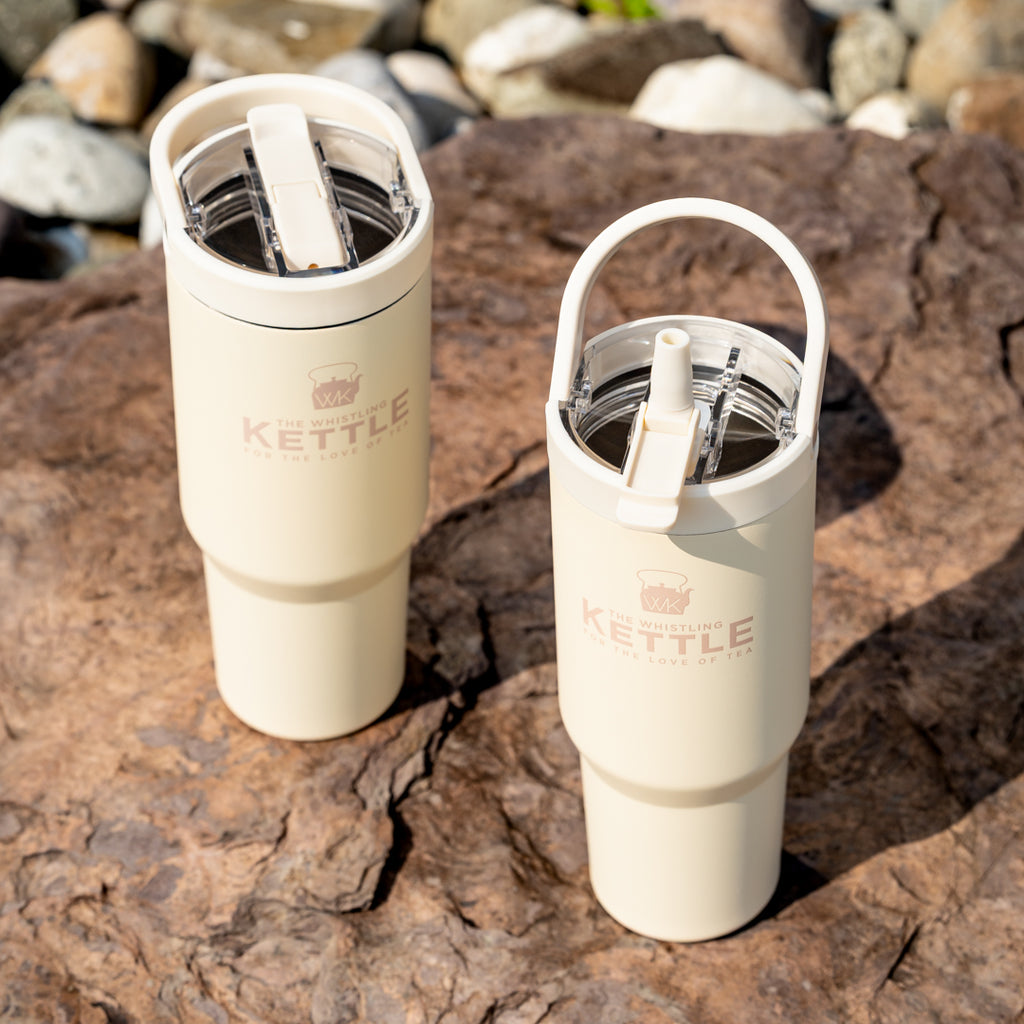 Pair of The Whistling Kettle Big Sipper insulated travel tumblers with carry handles and leak-resistant flip-top lids displayed on sunlit rocks