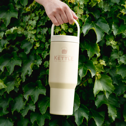 The Whistling Kettle Big Sipper insulated travel tumbler with a built-in carry handle held against a backdrop of green ivy