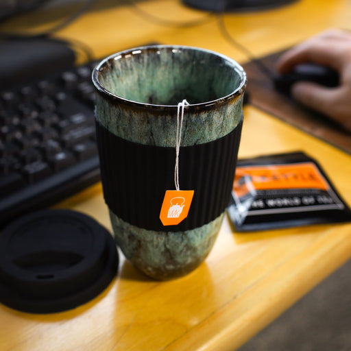 Green marbled ceramic travel mug with black silicone sleeve and tea bag on an office desk beside a keyboard and computer