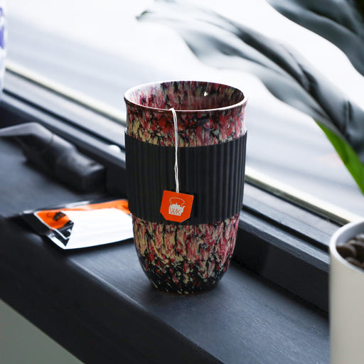 Red marbled ceramic travel mug with black silicone sleeve and tea bag on a windowsill next to a packet of tea