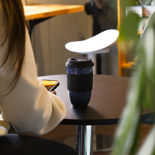Blue marbled ceramic travel mug with black silicone sleeve and lid on a café table beside a person using a smartphone