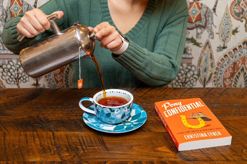 Pouring belgian chocolate tea into a cup and saucer next to Pony Confidential book