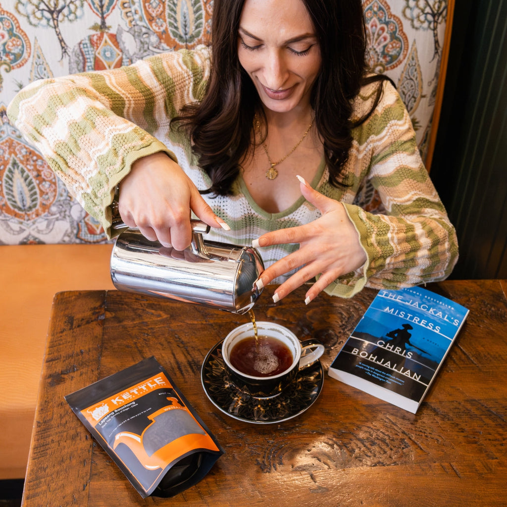 A woman pouring hot lapsang souchong tea into a cup with the jackal's mistress book