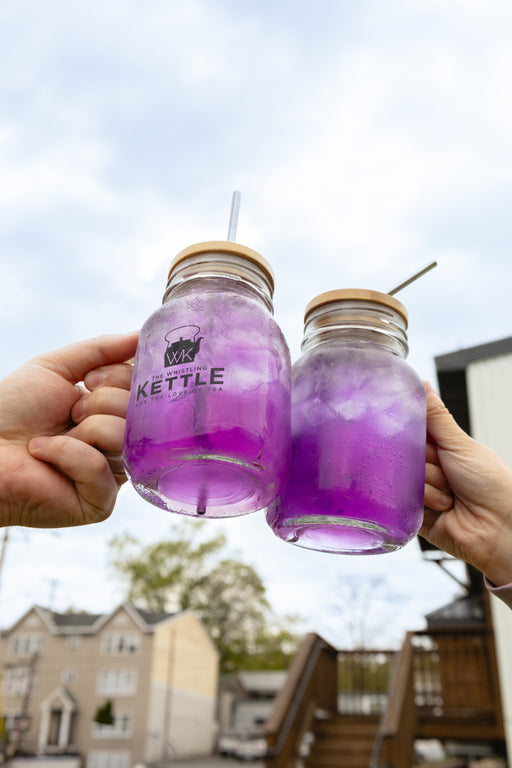 Two hands holding mason jars with purple iced tea against a blurred outdoor background.