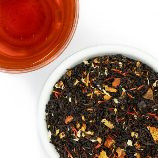 Overhead shot of a white bowl filled with loose-leaf herbal tea blend, with brewed amber-red tea visible in a glass cup at the top left