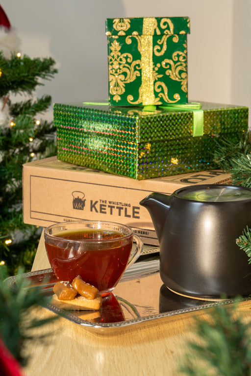A festive holiday scene featuring a stack of green patterned gift boxes, with a glass teacup of tea and a dark teapot on a tray with caramel bites on the side