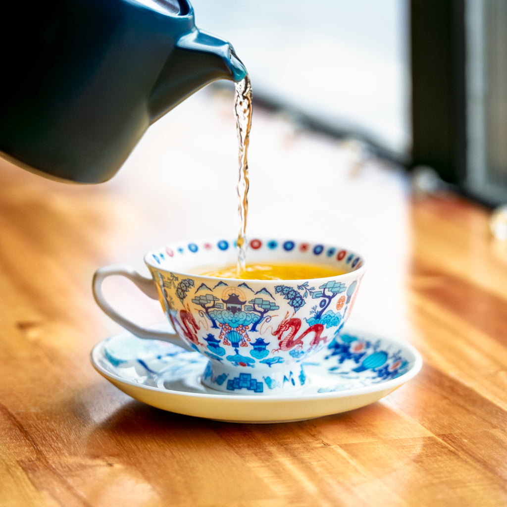 A slate-colored small teapot, partially off-screen, pours tea into a blue and red decorative Chinoiserie tea cup and saucer set