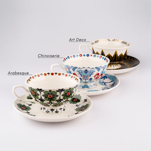 Three tea cup and saucer sets are lined up to showcase their various designs with a black font label for the name of each design, the first called Arabesque, the second called Chinoiserie, and the third called Art Deco