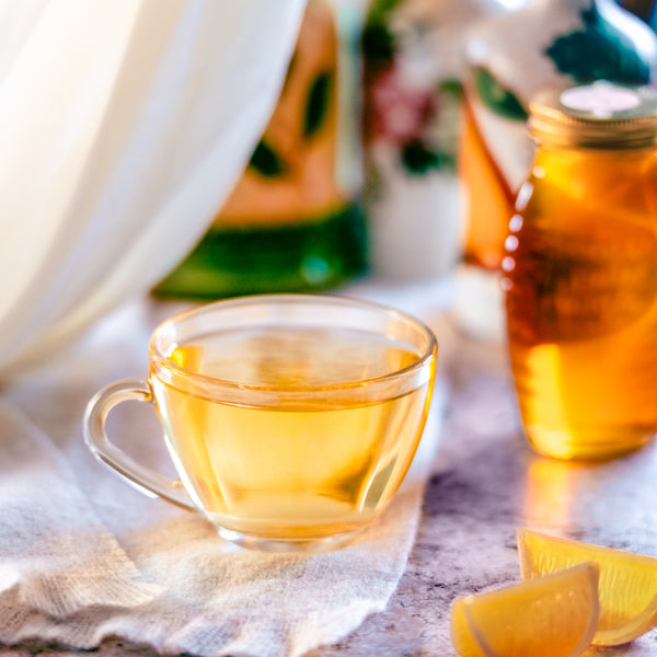 Glass cup of golden herbal tea with lemon wedges and a jar of brewed tea and a jar of honey blurred  in the background