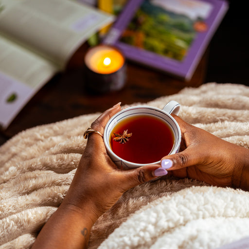 Person holding a cup of deep amber caramel apple chai tea while relaxing under a cream-colored blanket