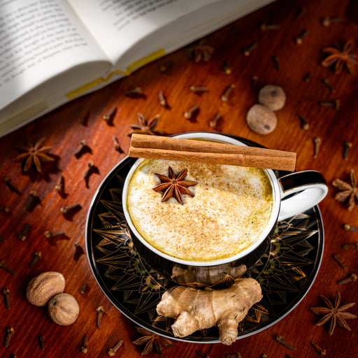 Overhead view of a frothy matcha chai latte topped with star anise and cinnamon sticks in a black teacup, surrounded by fresh ginger, cloves, nutmeg, star anise, and an open book on a wooden table