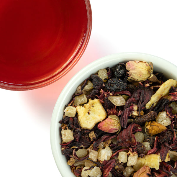 A deep red steeped cup of Cherry Bomb fruit tea next to a bowl of loose Cherry fruit tea containing sour cherries, pineapple cubes, hibiscus, sultanas, banana, and rosebuds