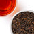 A top-down view showing a white bowl filled with dark loose-leaf tea leaves next to a clear glass cup of brewed amber-red tea on a white background