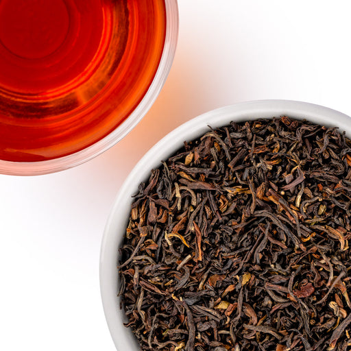 A top-down view showing a white bowl filled with dark loose-leaf tea leaves next to a clear glass cup of brewed amber-red tea on a white background