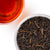 A top-down view showing a white bowl filled with dark loose-leaf tea and a glass of amber-red brewed tea on a white background