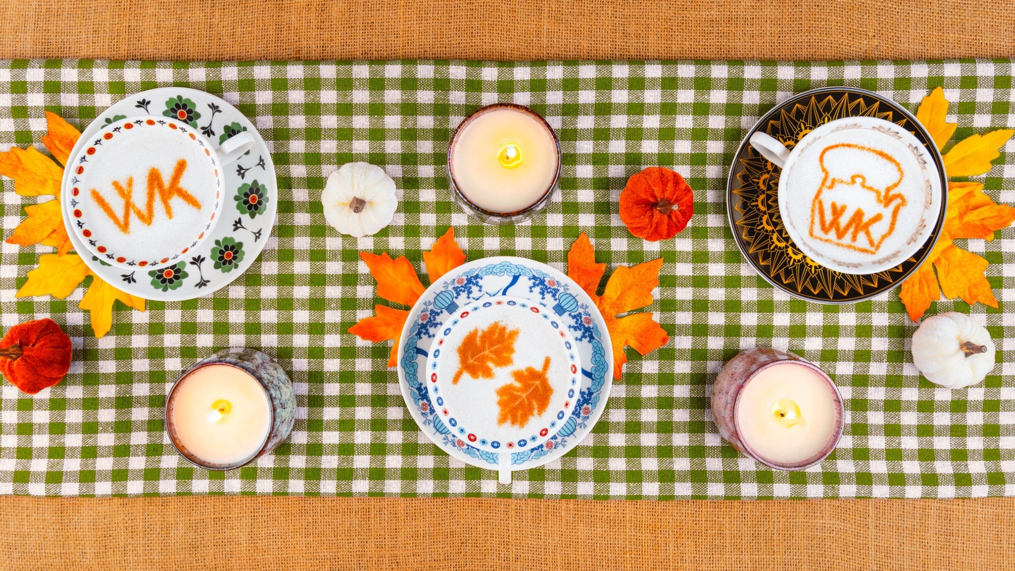 Top-down styled autumn table scene with a green-and-white checkered runner, burlap surface, candles, fall leaves, small pumpkins, and two decorated cups or mugs with latte art and monogram details.