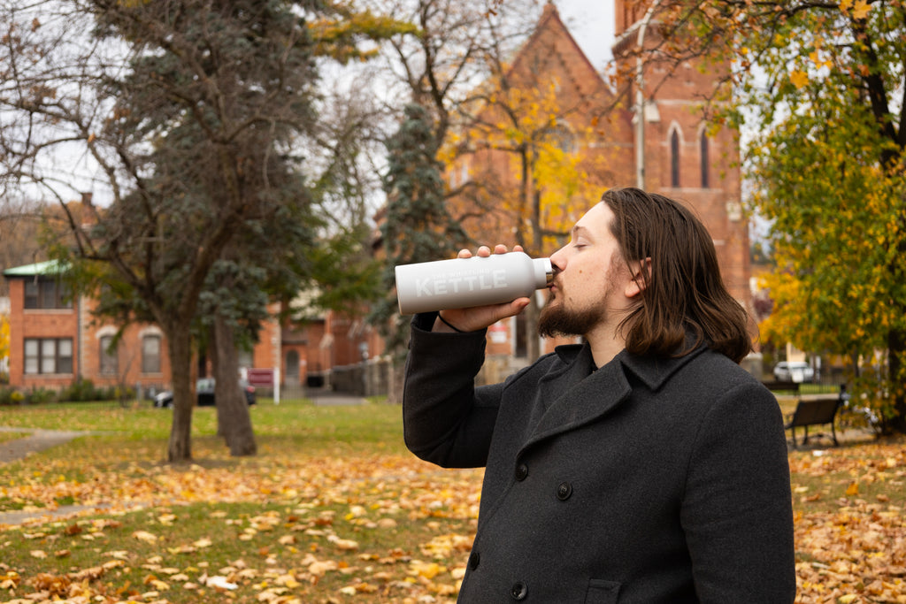 Man drinking from a bottle in an outdoor setting with trees and buildings in the background