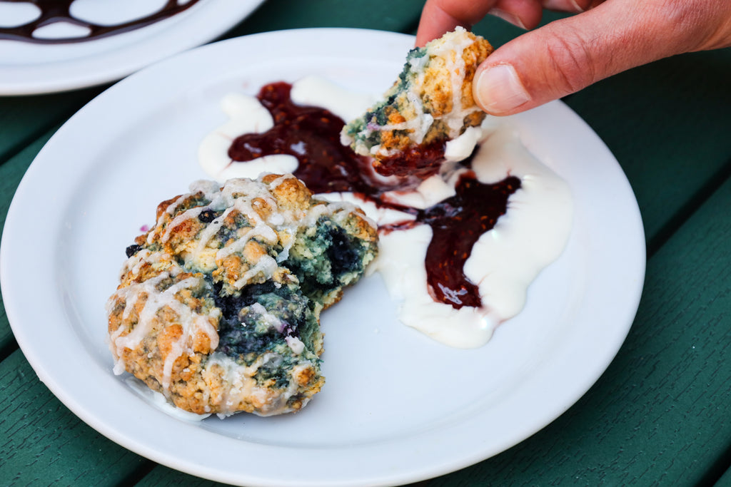 A close-up of a hand dipping a blueberry scone into cream cheese icing and berry jam atop a white plate
