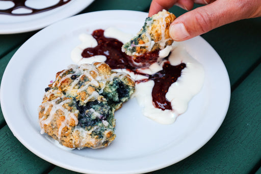 A close-up of a hand dipping a blueberry scone into cream cheese icing and berry jam atop a white plate