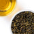 Overhead view of tightly rolled, dark green Gardenia Oolong tea leaves in a white bowl next to a cup of golden-amber brewed tea on a white background
