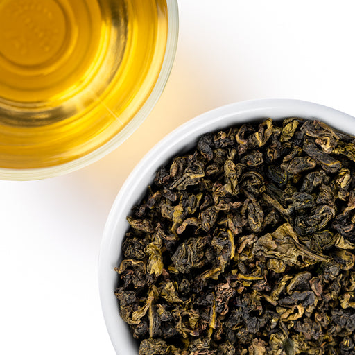 Overhead view of tightly rolled, dark green Gardenia Oolong tea leaves in a white bowl next to a cup of golden-amber brewed tea on a white background