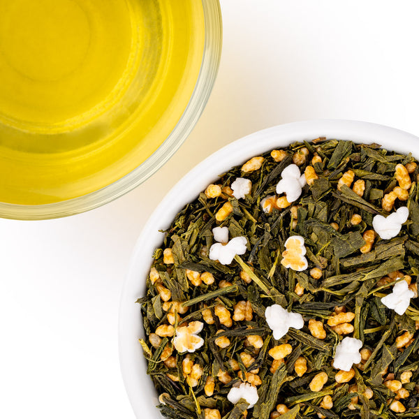 Overhead shot of loose-leaf tea with green leaves, golden-yellow flower buds, and white popcorn kernels in a white bowl. Next to the bowl is a glass of brewed yellow tea 