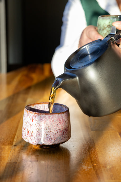 Tea being poured from a teapot into a ceramic cup on a wooden surface.