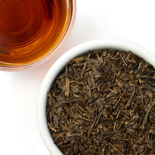 Dry tea leaves in a white bowl with a glass of brewed tea on a white background