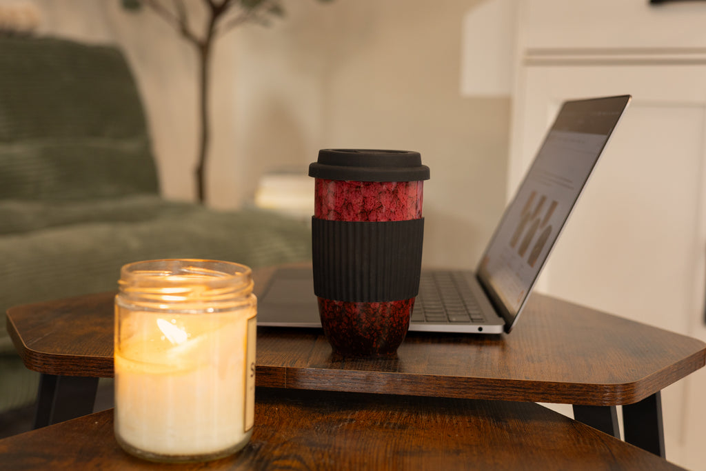 Red marbled ceramic travel mug with black silicone sleeve beside a laptop and lit candle on a wooden coffee table