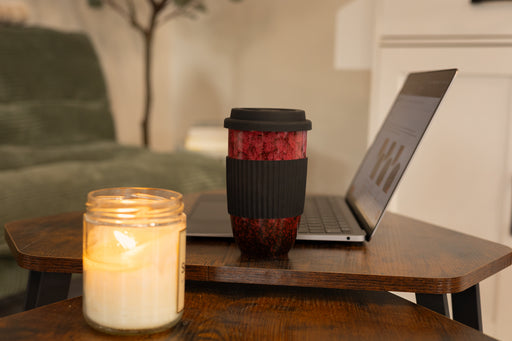 Red marbled ceramic travel mug with black silicone sleeve beside a laptop and lit candle on a wooden coffee table