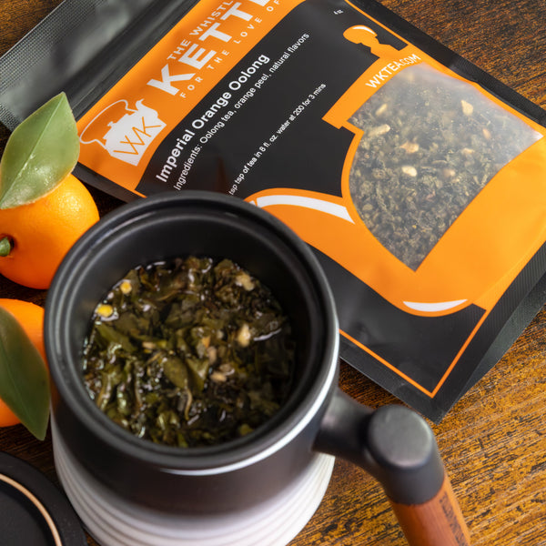 Mug with infuser steeping oolong tea leaves next to a 4oz size bag of Imperial Orange Oolong tea with oranges sitting next to the bag