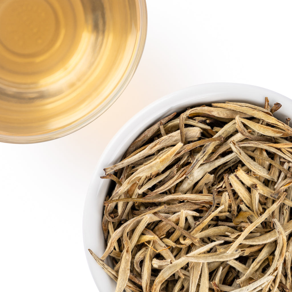Overhead view of Jasmine Silver Needles loose-leaf white tea in a white bowl beside a cup of pale golden brewed tea on a white background
