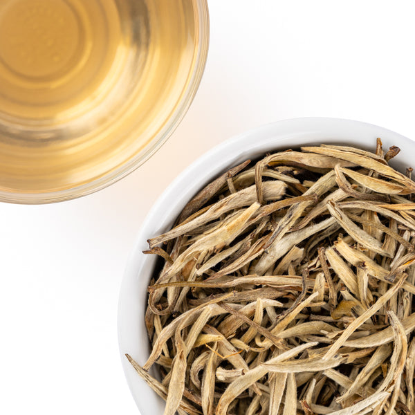 Overhead view of Jasmine Silver Needles loose-leaf white tea in a white bowl beside a cup of pale golden brewed tea on a white background