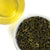 Dry green tea leaves in a white bowl with a glass of brewed tea on a white background