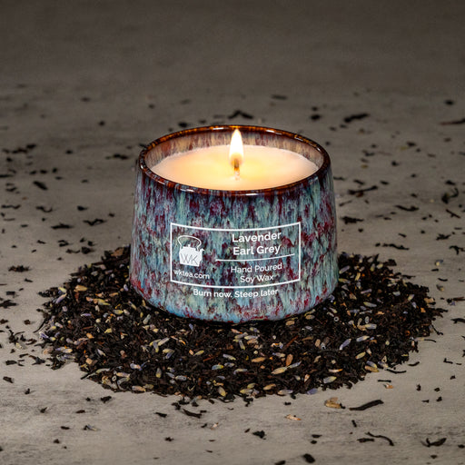 A lit candle in a textured blue and purple marbled ceramic tea cup, surrounded by a ring of black tea leaves and lavender petals