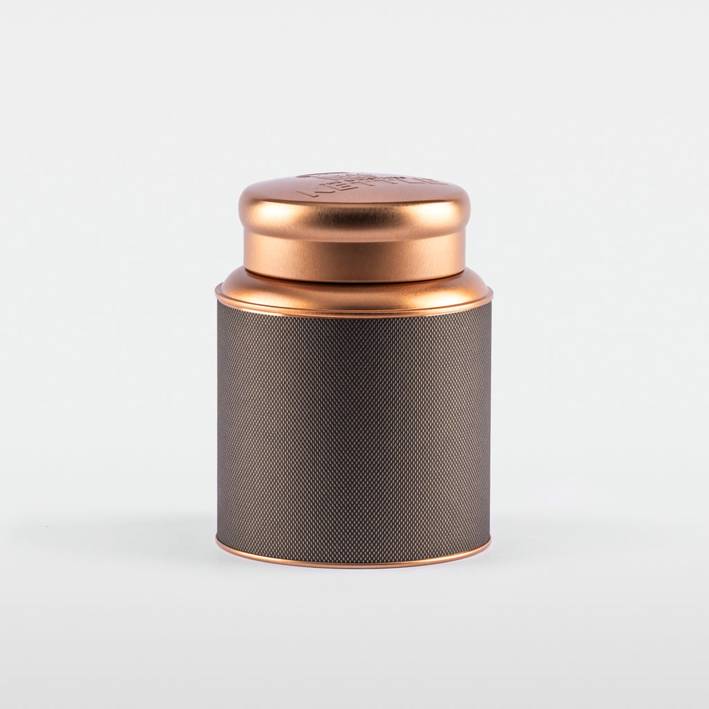 A cylindrical stainless steel tea tin with a textured black label and a shiny rose-gold lid