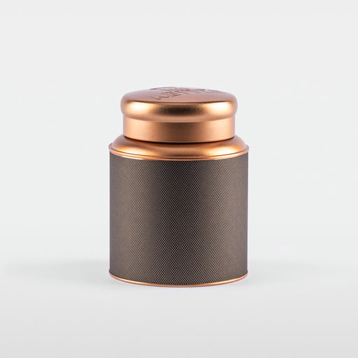 A cylindrical stainless steel tea tin with a textured black label and a shiny rose-gold lid