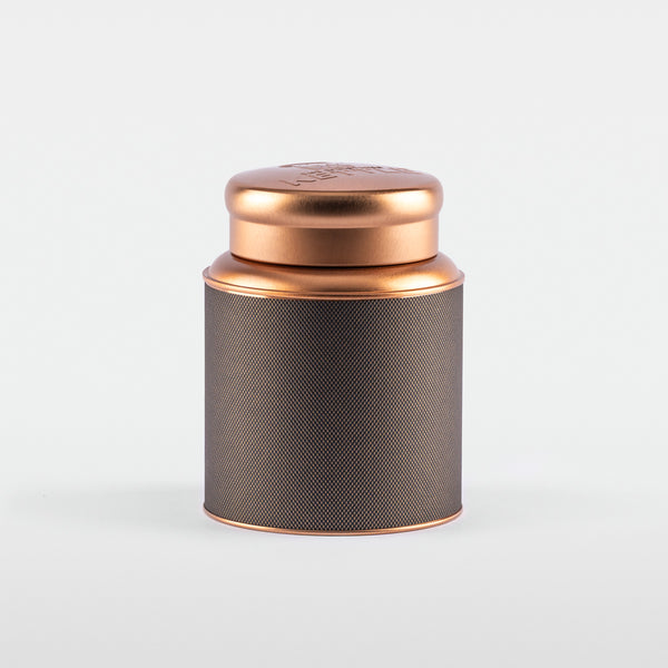 A cylindrical stainless steel tea tin with a textured black label and a shiny rose-gold lid