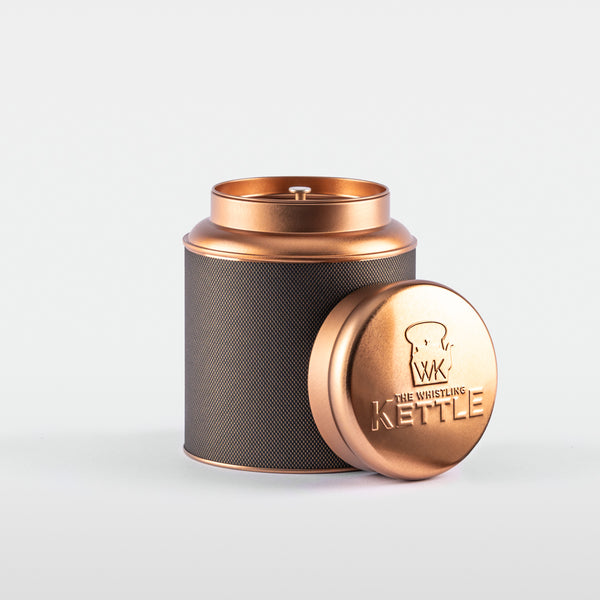 A cylindrical stainless steel tea tin with a textured black label and a shiny rose-gold lid