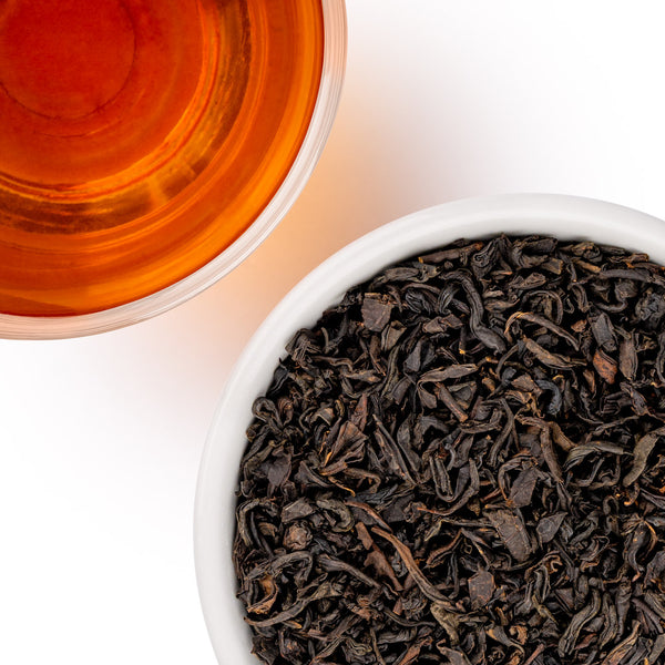 A close-up overhead shot of loose-leaf tea leaves in a white bowl next to a glass of amber tea on a white background.