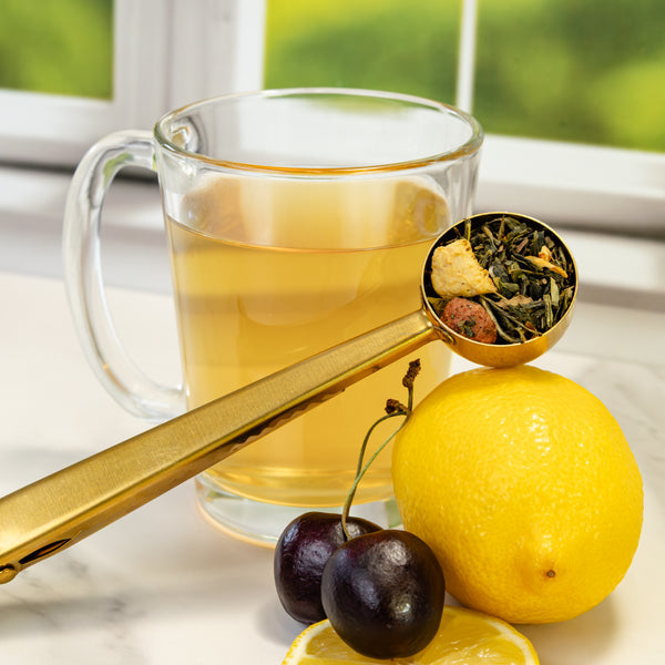 A yellow steeped cup of lemon cherry tea with lemon and cherries, next to a scoop of loose leaf green and white lemon cherry tea