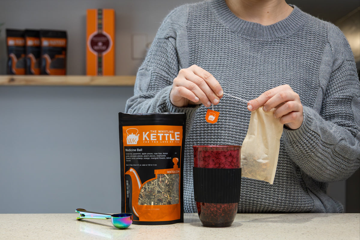 A person holding a tea bag standing at a kitchen counter. In front of them is a red ceramic bird's egg tea cup, a 4oz bag of Medicine Ball loose leaf tea, and a tea scoop.