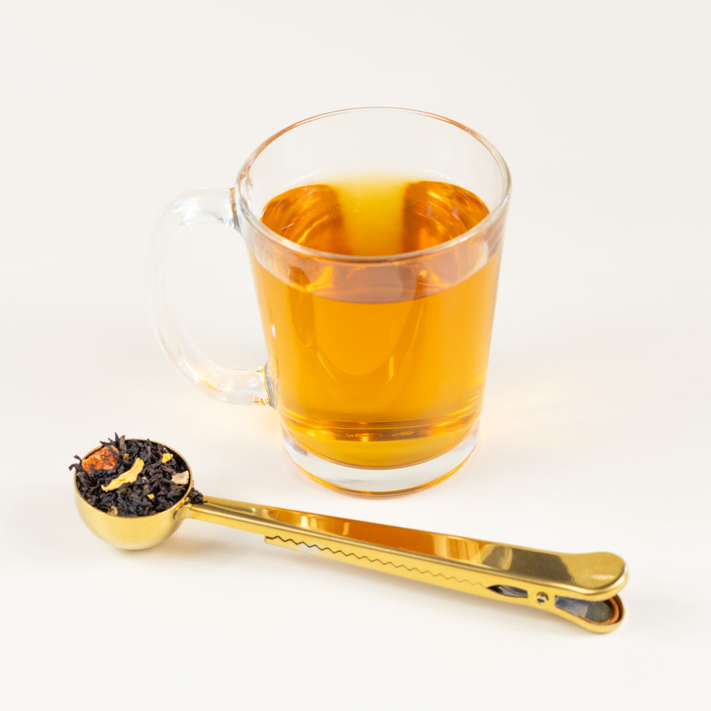 Glass of brewed Mango Passionfruit tea next to Gold Tea Scoop filled with Mango Passionfruit loose leaf tea. 